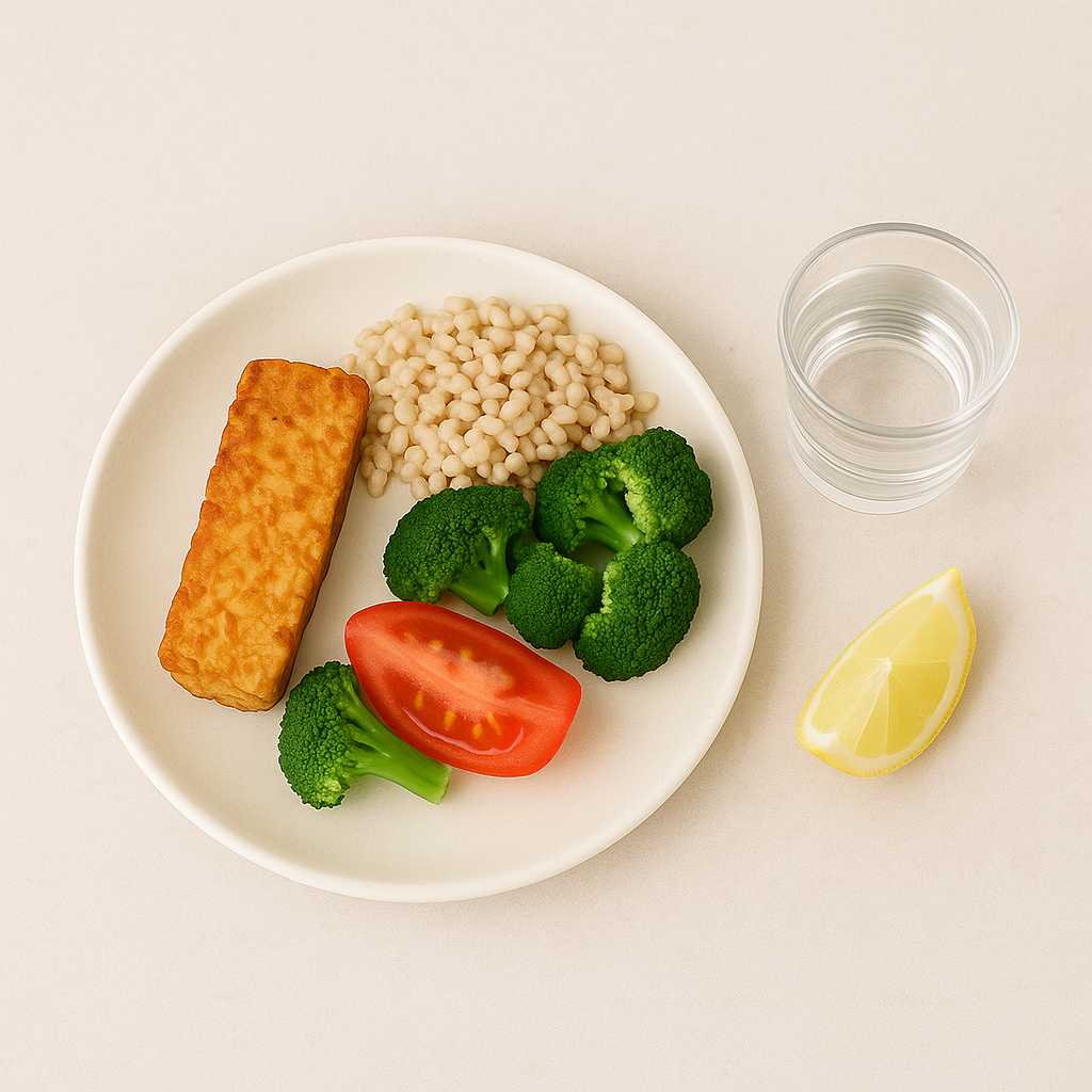 Plate of wholesome plant-based Panchakarma detox meal with grilled tempeh, barley, steamed broccoli, tomato slice, a glass of water, and lemon wedge.